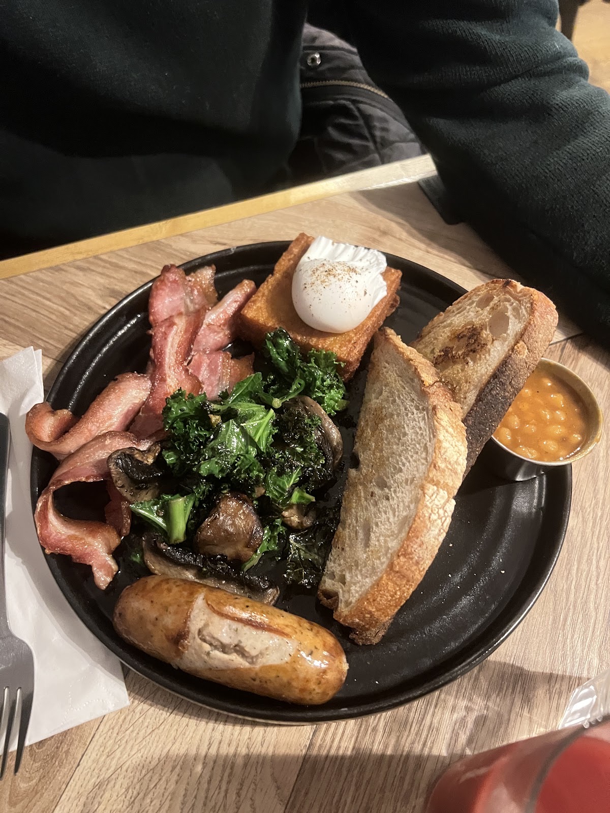 A full cooked breakfast in a black pan with bacon, poached egg, mushrooms, kale, sausage, beans and toast