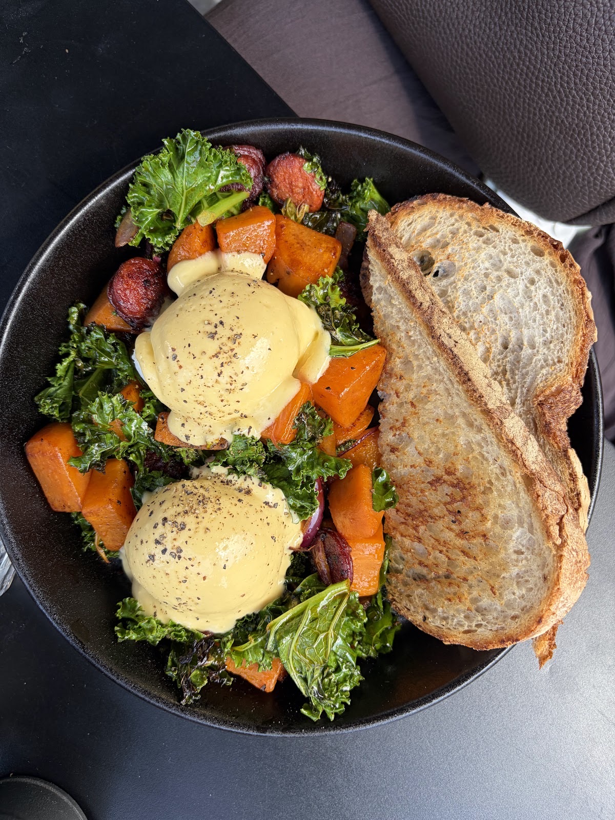 Eggs benedict with kale, roast squash and a halved toasted sandwich in a dark pan